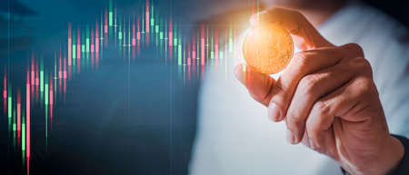 Businessman's hand holds the floating gold Bitcoin which is virtual money on a with a backdrop of the falling stock graph. Make money with crypto currency, blockchain transfer.の写真素材
