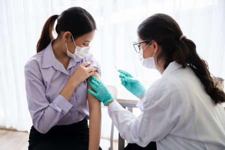 A female doctor or nurse wearing a mask, glove, and face shield are injecting the virus vaccine on the shoulder of the woman to immunize.の写真素材