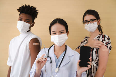 Female doctor stands holding a syringe and a smartphone, with a young man and woman various nationalities standing behind him wearing masks.の写真素材