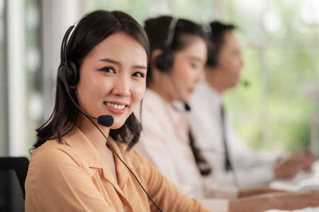 Team of call center staff in Asia wears headphones with a microphone. Smile while serving customers at desks and computers. Service concept and consulting. Communication concept.の写真素材