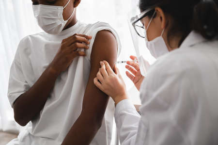 A female doctor or nurse wearing a mask and face shield is injecting the coronavirus 19 vaccine on the shoulder of an African-American man to immunize. Concept of preventing the spread of COVID-19.の写真素材