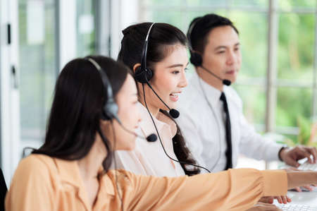 Team of call center staff in Asia wears headphones with a microphone. Smile while serving customers at desks and computers. Service concept and consulting. Communication concept.の写真素材