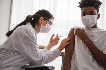 A female doctor or nurse wearing a mask and face shield is injecting the coronavirus 19 vaccine on the shoulder of an African-American man to immunize. Concept of preventing the spread of COVID-19.の写真素材