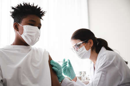 A female doctor or nurse wearing mask,glove and face shield is injecting the coronavirus 19 vaccine on the shoulder of African-American man to immunize. Concept of preventing the spread of COVID-19.の写真素材