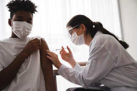 A female doctor or nurse wearing a mask and face shield is injecting the coronavirus 19 vaccine on the shoulder of an African-American man to immunize. Concept of preventing the spread of COVID-19.の写真素材