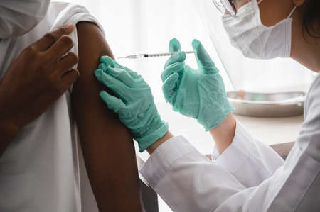 A female doctor or nurse wearing mask,glove and face shield is injecting the coronavirus 19 vaccine on the shoulder of African-American man to immunize. Concept of preventing the spread of COVID-19.の写真素材