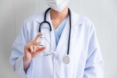 Female doctor wearing a mask stands holds a bottle of vaccine for protection Coronavirus19 in a laboratory. Concept of preventing the spread of COVID-19.の写真素材