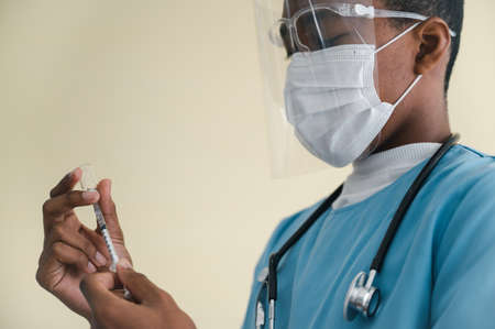 African-American man doctor wearing a mask and face shield. Holding a syringe with a bottle of coronavirus19 vaccine protection in a laboratory. Concept of protection against the COVID-19 virus.の写真素材