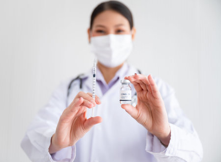 Female doctor wearing a mask stands holding a syringe with a vaccine. Coronavirus19 in the laboratory. Concept of protection against the COVID-19 virus.の写真素材