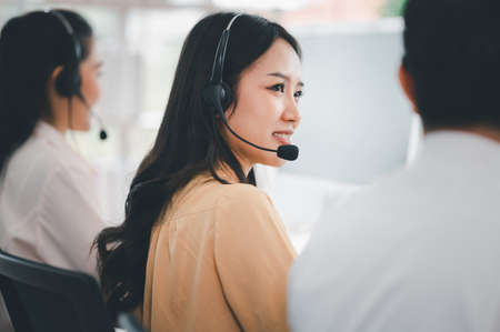Team of call center staff in Asia wears headphones with a microphone. Smile while serving customers at desks and computers. Service concept and consulting. Communication concept.の写真素材