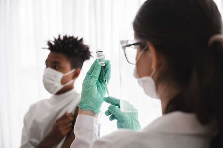Female doctor or nurse wearing mask, gloves, and visor preparing to vaccinate coronavirus 19 to immunize African-American men.Concepts to prevent the spread of COVID-19.の写真素材