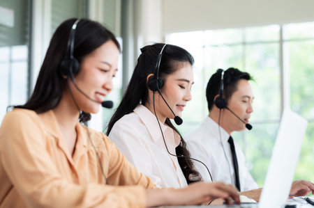 Team of call center staff in Asia wears headphones with a microphone. Smile while serving customers at desks and computers. Service concept and consulting. Communication concept.の写真素材