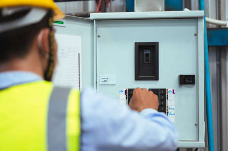 Engineer, Supervisor or Foreman male wear a mask, hard hat, and vest. Checking the power control box panel and electrical system inside the factory. for safety at work.の写真素材