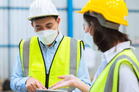 Team engineers and foreman wear a mask, hard hat, and vest. Standing consult discuss industrial production management. Explaining job details through laptops and tablets in factories or warehouses.の写真素材