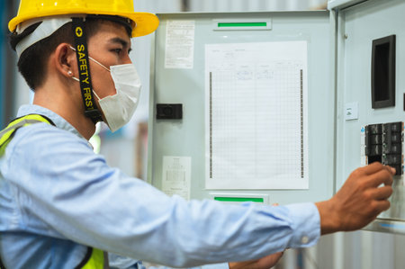 Engineer, Supervisor or Foreman male wear a mask, hard hat, and vest. Checking the power control box panel and electrical system inside the factory. for safety at work.の写真素材