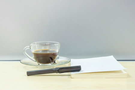 Closeup black pen on white book with black coffee in transparent cup of coffee on blurred wooden desk and frosted glass wall textured backgroundの写真素材
