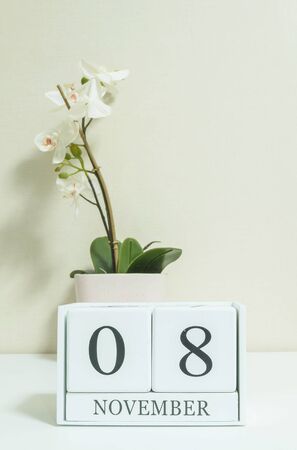 Closeup white wooden calendar with black 8 november word with white orchid flower on white wood desk and cream color wallpaper in room textured background , selective focus at the calendarの写真素材