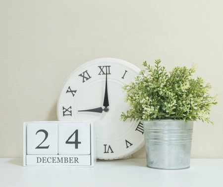 White wooden calendar with black 24 december word with clock and plant on white wood desk and cream...
