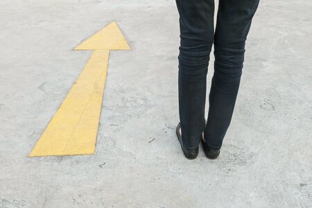 Closeup woman foot in black trousers with old and pale yellow painted arrow sign on cement street floor textured backgroundの写真素材