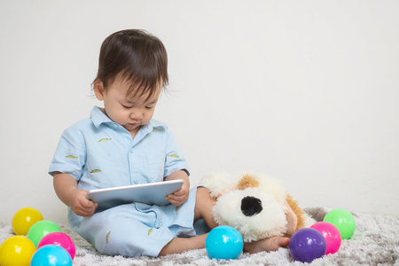 Closeup cute asian kid look at the tablet at home on gray carpet with doll and colorful ball and cement wall textured background with copy spaceの写真素材