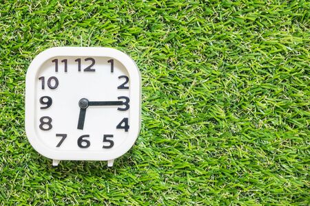 Closeup white clock for decorate show a quarter past six or 6:15 a.m. on green artificial grass floor textured background with copy spaceの写真素材