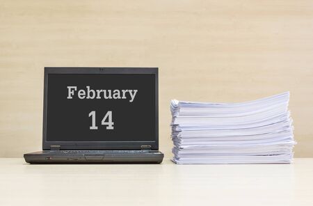 Closeup computer laptop with february 14 word on the center of screen in calendar concept and pile of work paper on wood desk and wood wall in work room textured background with copy spaceの写真素材