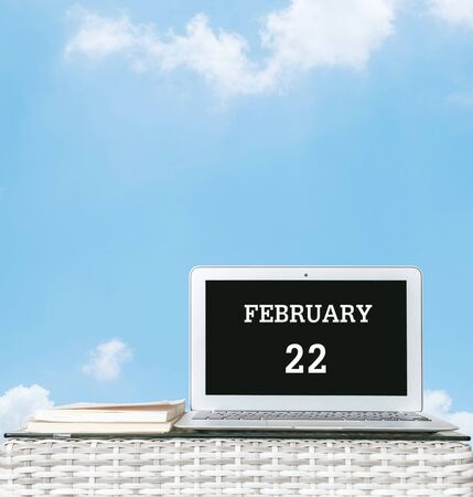 Closeup computer laptop with february 22 word on the center of screen in calendar concept on blurred wood weave table and book on blue sky with cloud textured background with copy spaceの写真素材