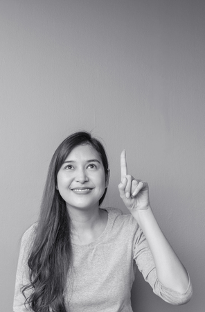 Closeup asian woman holds up one finger point to space with smile face on blurred cement wall textured background in black and white tone with copy spaceの写真素材