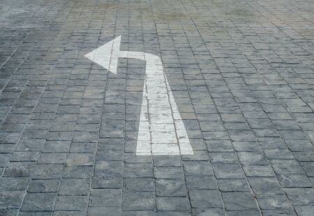 Closeup white painted arrow sign in left side on black brick stone floor at the car park textured backgroundの写真素材