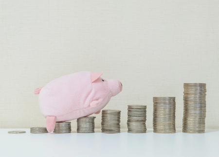 Closeup pile of coin start from low to high with pig doll on white wood desk and wallpaper textured background with copy space in saving money conceptの写真素材