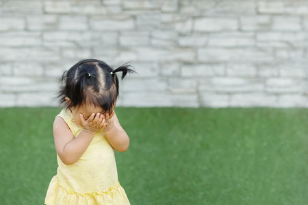 Closeup asian girl take her hands off the face and play hidden with someone on grass floor and brick wall textured background with copy spaceの写真素材