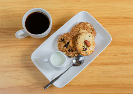 Black Coffee, Almond Cookies and Cashew cookies in white plate and fresh milk in white cup on brown wooden background.の写真素材