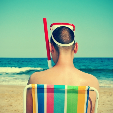 picture from the back of a man wearing a diving mask and a snorkel seated in a deckchair on the beach, with a retro effectの写真素材