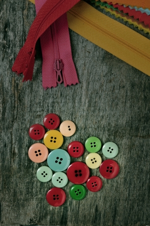 a pile of buttons of different colors forming a heart in an old wooden background with zippersの写真素材