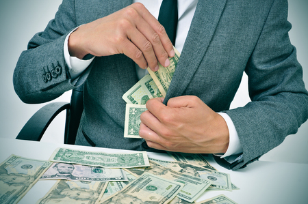 man in suit sitting in a desk full of dollar bills getting them in his jacketの写真素材