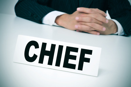 a man wearing a suit sitting in a desk with a signboard in front of him with the word chief written in itの写真素材