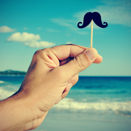 a man hand holding a fake moustache in a stick in his hand on the beach, with a filter effectの写真素材