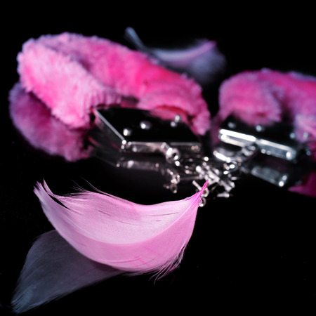 a pair of pink sexy fluffy handcuffs and some pink feathers used as adult toys on a reflecting black backgroundの写真素材