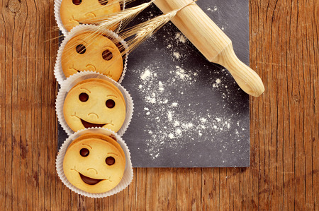 high-angle shot of some smiley biscuits, some ears of wheat, a wooden rolling pin, and wheat flour sprinkled in a black slate plate placed on a wooden rustic tableの写真素材