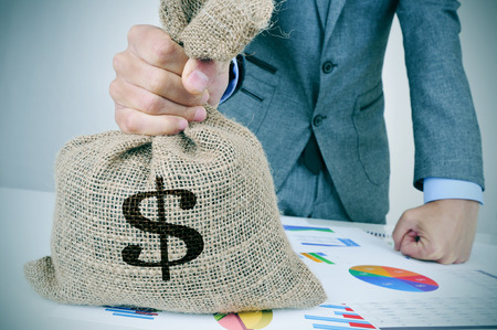 closeup of a young caucasian man in a grey suit with a money bag on a desk full of different chartsの写真素材