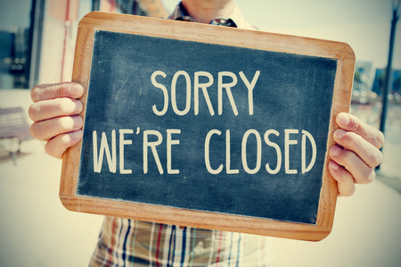 closeup of a young caucasian man wearing a plaid shirt shows a chalkboard with the text sorry we are closed written in it, with a filter effectの写真素材