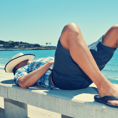 a young caucasian man with a straw hat on his face lying down in a street bench near the seaの写真素材