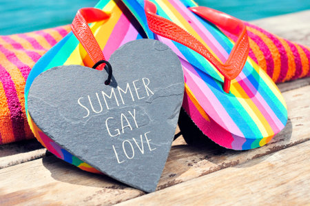 the sentence summer gay love written in a heart-shaped slate stone and a pair of rainbow flip-flops and a beach towel on a boardwalk above the oceanの写真素材