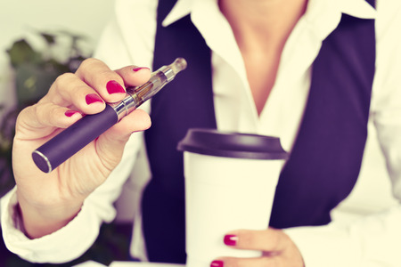 a young caucasian woman, with a cup of coffee in her hand, vapes from an electronic cigaretteの写真素材