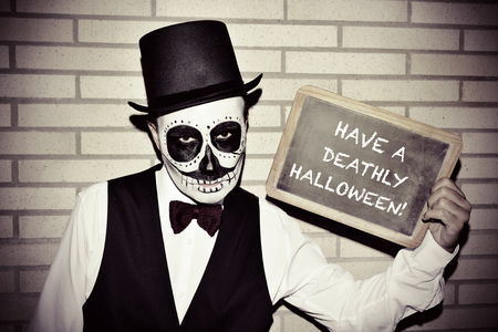 a man with calaveras makeup, wearing bow tie and top hat, against a brick wall, shows a chalkboard with text have a deathly halloween written in itの写真素材