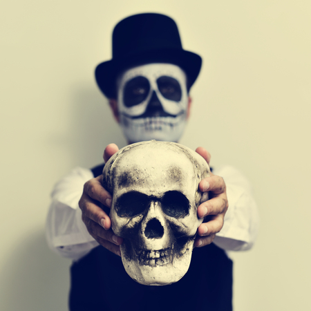 a young man with calaveras makeup, wearing top hat, holds a scary skull in front of himの写真素材