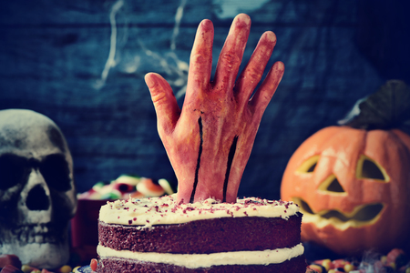 a red velvet cake topped with a bloody hand in a scary scene for halloween with a carved pumpkin, a skull and cobwebsの写真素材