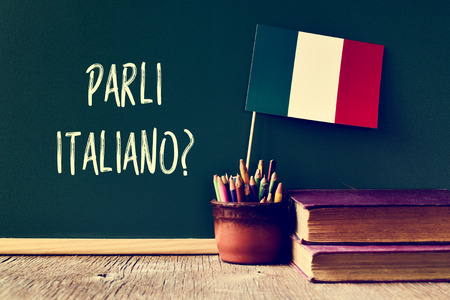 a chalkboard with the question parli italiano? do you speak Italian? written in Italian, a pot with pencils, some books and the flag of Italy, on a wooden deskの写真素材