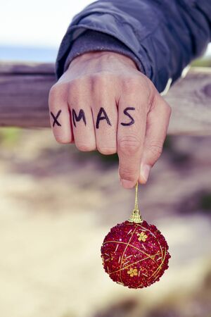 closeup of a young caucasian man with the word xmas written in the knuckles of his hand holding a red christmas ball outdoorsの写真素材