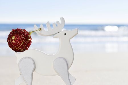 closeup of a red christmas ball in the antler of a white wooden reindeer, with the sea in the backgroundの写真素材